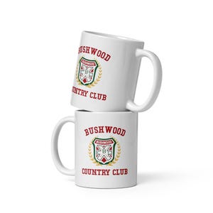 May include: Two white ceramic mugs stacked on top of each other. Each mug features the words "BUSHWOOD COUNTRY CLUB" in red lettering, with a crest design in the center. The crest includes the word "BUSHWOOD" and a geometric design.