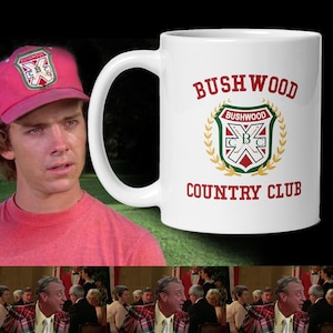 May include: White ceramic mug with the text "Bushwood Country Club" in red lettering. The mug features a crest design with the words "Bushwood" and "CBC" inside a shield, surrounded by a green and gold wreath. A pink baseball cap with a similar crest is also visible.