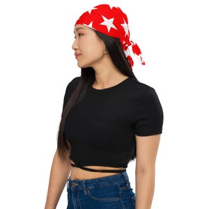 May include: A woman wearing a red bandana with white stars. The bandana is tied around her head and has a knot at the side. She is wearing a black crop top and blue jeans.