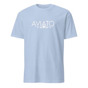 Puede incluir: Camiseta azul claro con la palabra "AVIATO" en blanco, sobre un gráfico de avión blanco. La camiseta tiene cuello redondo y mangas cortas. La tela parece suave y cómoda.
