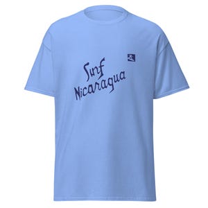 Puede incluir: Camiseta azul claro con las palabras "Surf Nicaragua" en escritura azul oscuro. Un pequeño gráfico azul oscuro de un surfista está en el lado derecho. La camiseta tiene mangas cortas y cuello redondo.