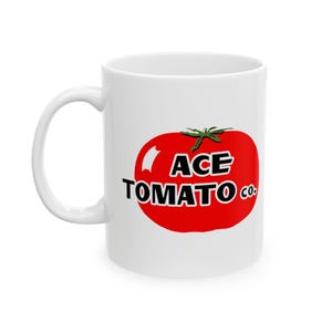 May include: White ceramic mug featuring a large red tomato graphic with the text "ACE TOMATO CO." in black. The mug has a curved handle and is designed for hot beverages. The design is simple and bold.