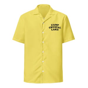 May include: A yellow short-sleeved button-down shirt with a camp collar. The shirt features the text "CAMP CRYSTAL LAKE" in black block letters on the front. The shirt has a classic, casual style.