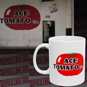 May include: White ceramic mug featuring a red tomato graphic with the text "ACE TOMATO CO." The background includes a wall with a large tomato graphic and the text "ACE TOMATO CO."