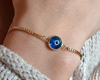 Può includere: Un braccialetto a catena dorata con un ciondolo blu a forma di occhio turco. Il ciondolo ha una pupilla bianca e un bordo dorato.