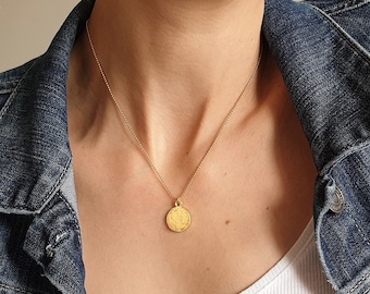 May include: A gold chain necklace with a round gold pendant. The necklace is worn by a person wearing a white tank top and a blue denim jacket.