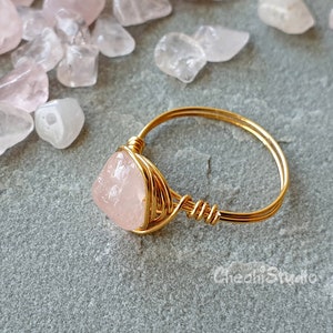 May include: A gold wire wrapped ring featuring a pink rose quartz stone. The ring is surrounded by small pink rose quartz chips.