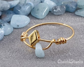 Puede incluir: Un anillo de oro con un pequeño dije cuadrado de oro. El anillo está rodeado de piedras de color azul claro.