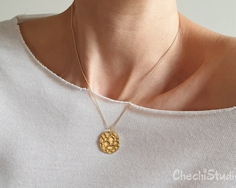May include: A gold chain necklace with a textured round pendant. The necklace is worn by a person wearing a white t-shirt.
