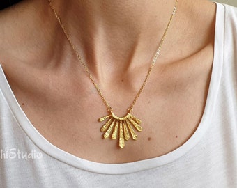 May include: A gold necklace with a delicate chain and a geometric pendant that resembles a sunburst. The pendant is made of multiple thin, textured pieces of metal that radiate outwards from a central point.