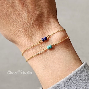 May include: Two delicate gold chain bracelets. One features two dark blue beads, the other has two turquoise beads. The bracelets are displayed on a wrist, showcasing the minimalist design and colour contrast.