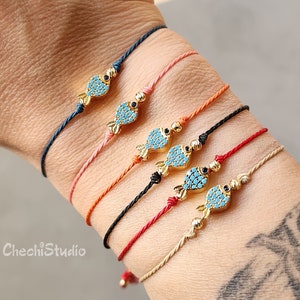 May include: Six colourful string bracelets with gold fish charms. The fish charms are decorated with turquoise and black enamel and have black eyes. The bracelets are tied in a knot on the wrist.