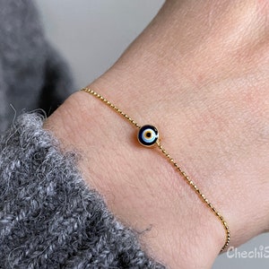 May include: A gold chain bracelet with a small blue evil eye charm. The charm has a white centre and a black outline.