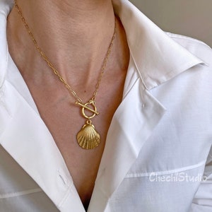Op de afbeelding: Een gouden ketting met een schelp hanger en een toggle sluiting. De ketting wordt gedragen door een model dat een wit shirt draagt.