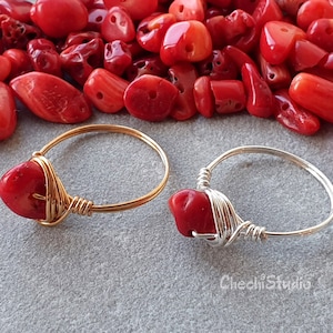 Puede incluir: Dos anillos con piedras de coral rojo envueltos en alambre dorado y plateado. Los anillos est&aacute;n sobre una superficie gris con cuentas de coral rojo esparcidas a su alrededor.