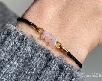 May include: A black cord bracelet with gold accents and three pink rose quartz stones.