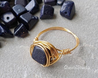 May include: Gold wire wrapped ring with a dark blue gemstone. The gemstone has a speckled appearance. The ring is surrounded by loose dark blue gemstones.
