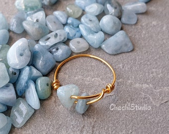 Puede incluir: Un anillo de alambre dorado envuelto con dos piedras de aguamarina azul claro. El anillo está rodeado de una dispersión de piedras de aguamarina azul claro.