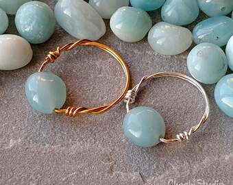 Puede incluir: Dos anillos envueltos en alambre con cuentas de piedras preciosas de color azul claro. El anillo de la izquierda está envuelto en alambre dorado y el anillo de la derecha está envuelto en alambre plateado.