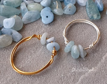 Puede incluir: Dos anillos de alambre con piedras de aguamarina envueltas alrededor. El anillo de la izquierda es de oro y el anillo de la derecha es de plata.