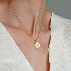 May include: A gold chain necklace with a round, textured pendant. The pendant has a detailed design and is worn against a white background.