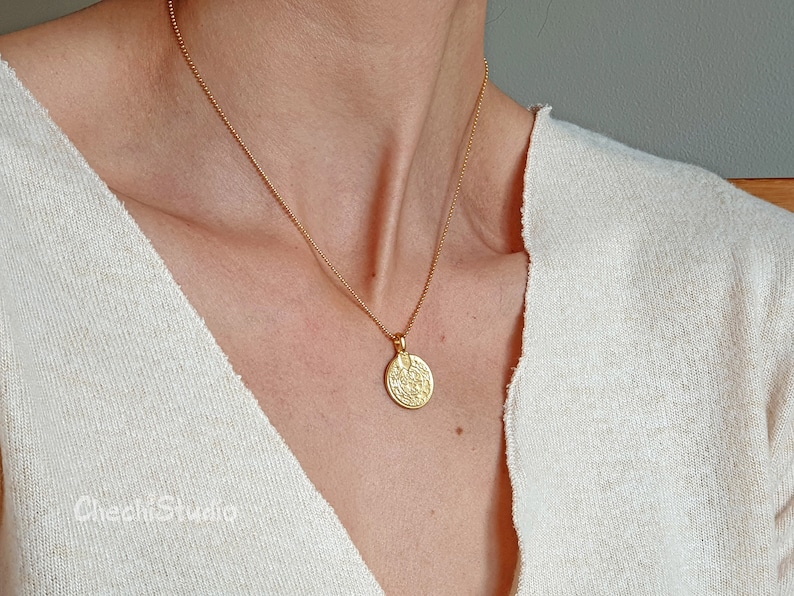 May include: A gold chain necklace with a round, textured pendant. The pendant has a detailed design and is worn against a white background.
