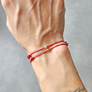 May include: Two red string bracelets, each with a small metallic bead. One bead is silver, the other is gold. The bracelets are tied with knots and worn on a wrist.