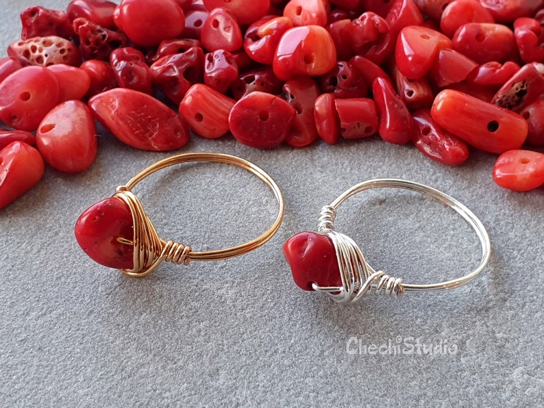 Puede incluir: Dos anillos con piedras de coral rojo envueltos en alambre dorado y plateado. Los anillos est&aacute;n sobre una superficie gris con cuentas de coral rojo esparcidas a su alrededor.