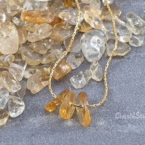 May include: A gold chain necklace featuring three teardrop-shaped citrine stones. The stones are a light golden yellow colour and are set in a simple, elegant design. The necklace is surrounded by a scattering of smaller, clear quartz stones.