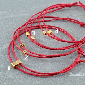 May include: Five red string bracelets with gold beads and charms. The bracelets are layered on top of each other. The charms are numbered 1 through 4.