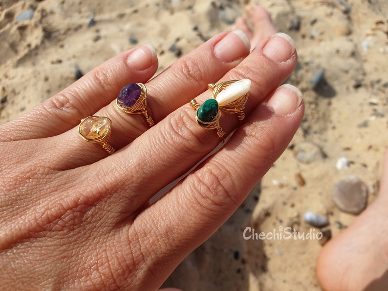 Puede incluir: Cuatro anillos de alambre dorado envueltos con diferentes piedras. Los anillos presentan una amatista p&uacute;rpura, un citrino amarillo, una malaquita verde y una concha blanca.