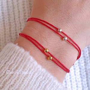 Puede incluir: Dos pulseras de cuerda roja con cuentas doradas y plateadas. Las pulseras se llevan en la muñeca.