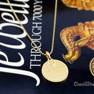 May include: A gold chain necklace with a round gold pendant featuring an intricate design. The pendant is hanging on a book with the text "THROUGH 7000 YEARS" visible.