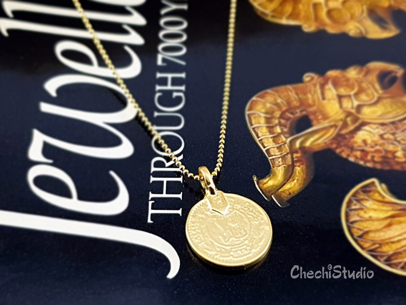May include: A gold chain necklace with a round gold pendant featuring an intricate design. The pendant is hanging on a book with the text "THROUGH 7000 YEARS" visible.