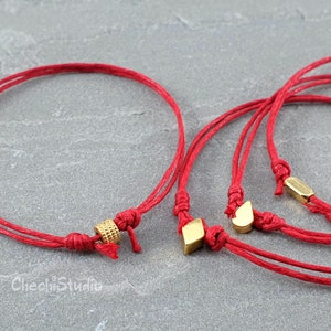 Puede incluir: Tres pulseras de cuerda roja con dijes de oro. Los dijes son un círculo pequeño, una forma de lágrima y una forma cuadrada.