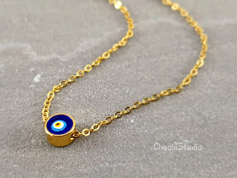 May include: A gold chain necklace with a small blue evil eye charm. The charm has a white center and a blue ring around it.