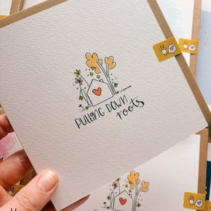 May include: A stack of greeting cards featuring a hand-drawn illustration of a house with a heart, trees, and the words "Pulling Down Roots." The cards have a kraft paper border. Small yellow stickers with a bird design are visible.