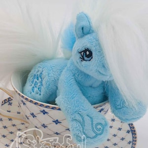May include: A light blue plush pony with a white mane and tail, featuring embroidered blue details on its flank and hoof, sitting inside a white teacup and saucer decorated with a blue and gold diamond pattern and the text Starful Stitches.
