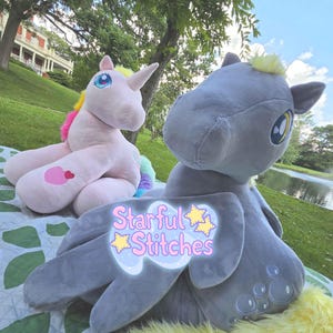 May include: Two plush toys, one light pink unicorn with a yellow and pink mane and a heart patch, and one grey winged creature with a yellow mane and bubble embroidery. A logo with the text Starful Stitches is centered over the grey plush toy.