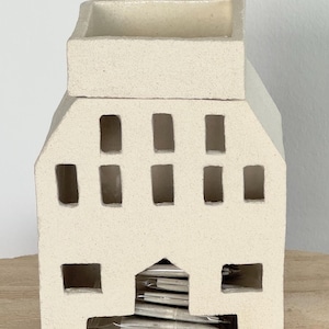 Può includere: Un portacandele a forma di casa in ceramica color crema con ritagli rettangolari per le finestre e un'apertura quadrata alla base. La parte superiore ha un'apertura rettangolare. All'interno dell'apertura della base sono visibili lumini.