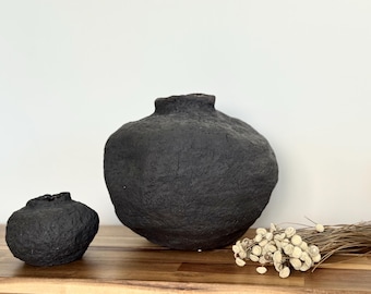 Op de afbeelding: Twee zwarte, getextureerde keramische vazen van verschillende grootte staan op een houten oppervlak. De grotere vaas heeft een afgeronde vorm met een smalle hals. Een kleinere, vergelijkbare vaas staat ernaast. Gedroogde bloemen zijn rechts.