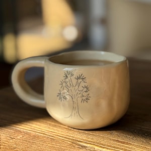 May include: Cream-colored ceramic mug with a rounded body and a large handle. A simple black tree design is on the front. The mug is filled with a dark liquid, possibly coffee or tea. The mug sits on a wooden surface.