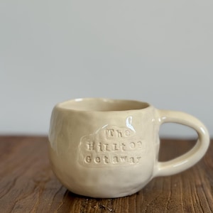 May include: Cream-coloured ceramic mug with a rounded shape and a handle. The mug features the embossed text "The Hilton Getaway" on its side. The mug is sitting on a wooden surface.