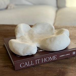 May include: A cream-colored, abstract decorative bowl with a textured surface, resting on a closed book. The book has the title "CALL IT HOME" in white lettering. The background includes a white sofa and a wooden table.