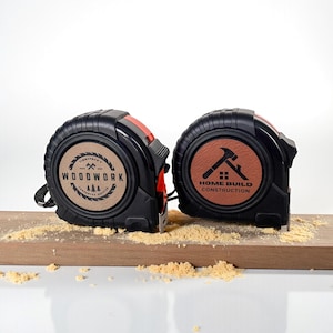 May include: Two black tape measures with red accents and custom logos. One reads "WOODWORK" and the other "HOME BUILD CONSTRUCTION". The tape measures are resting on a wooden surface with sawdust.