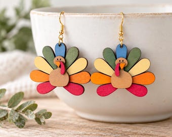 May include: A pair of dangle earrings featuring a turkey design made from layered wood pieces. The turkey has a tan body, a red wattle, and a fan of tail feathers in shades of green, yellow, orange, and red. The earrings hang from gold-toned hooks.
