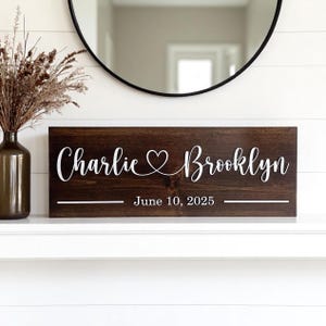 May include: A dark brown wooden sign with white lettering. The sign reads "Charlie &hearts; Brooklyn" with the date "June 10, 2025" below. The sign is rectangular and has a rustic, farmhouse style.