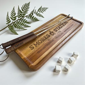 May include: A wooden S'mores Station serving tray with two roasting forks and marshmallows. The tray has the words "S'Mores Station" and "Graham Crackers - Chocolate - Marshmallows" engraved on it. The roasting forks have wooden handles with the names "Brian" and "Sarah" engraved on them.