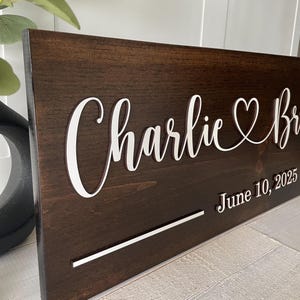 May include: A dark brown wooden sign with white lettering. The sign reads "Charlie & Bri" with a heart symbol and "June 10, 2025". A white horizontal line is below the names. The sign is likely for a wedding or anniversary.
