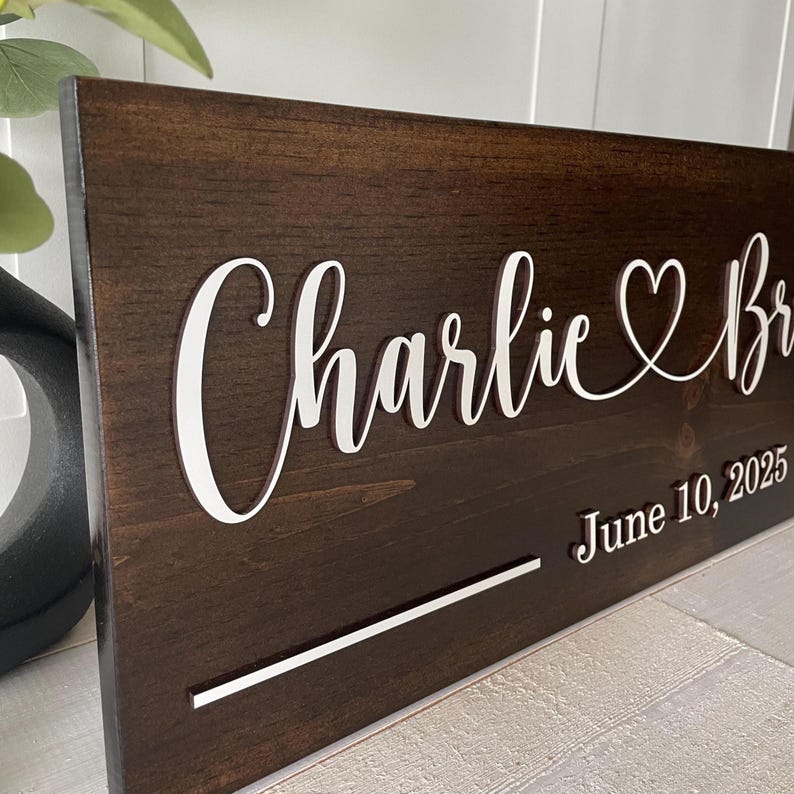 May include: A dark brown wooden sign with white lettering. The sign reads "Charlie & Bri" with a heart symbol and "June 10, 2025". A white horizontal line is below the names. The sign is likely for a wedding or anniversary.